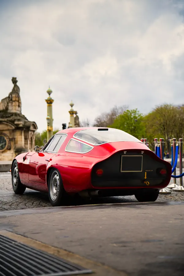 Alfa Romeo Giulia TZ rouge photographiée de trois-quarts arrière, montrant sa silhouette aérodynamique avec lunette arrière fastback et partie arrière tronquée, avec les colonnes rostrales et une statue en arrière-plan sous un ciel nuageux.