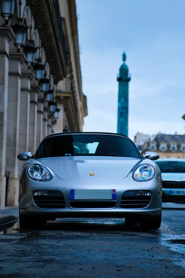 Porsche Boxster gris argenté stationnée sur une rue pavée humide de Paris, photographiée de face avec la colonne Vendôme visible en arrière-plan.