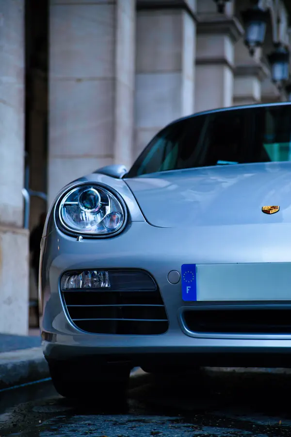 Gros plan sur l'avant d'une Porsche Boxster gris argenté montrant le phare ovale caractéristique, l'écusson Porsche sur l'aile, la plaque d'immatriculation française et une partie du pare-chocs avec prises d'air, stationnée devant une façade en pierre de taille.