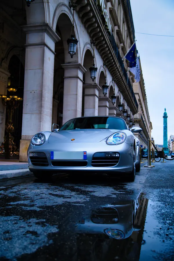 Porsche Boxster argentée garée devant un bâtiment historique parisien à l'architecture classique avec colonnes et lanternes, reflétée dans une flaque d'eau sur le pavé humide, avec la place Vendôme visible en arrière-plan.