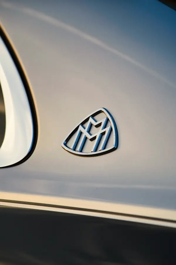 Logo Maybach en forme d'écusson triangulaire chromé sur la carrosserie blanche d'une voiture de luxe, photographié en gros plan avec une lumière douce mettant en valeur les contours métalliques du badge.