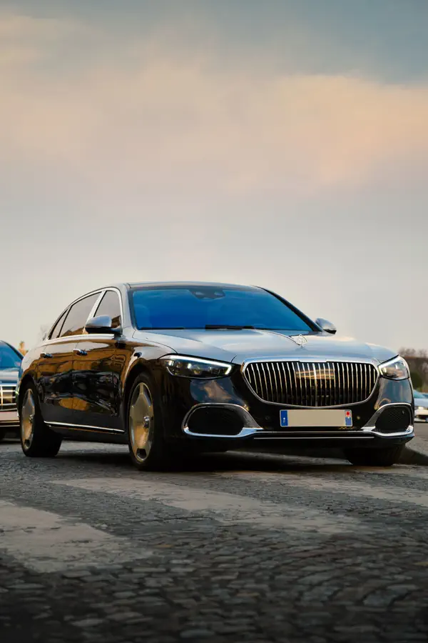 Mercedes-Maybach Classe S noire photographiée sur une surface pavée, vue de trois-quarts avant, avec sa calandre verticale chromée imposante, son emblème Maybach sur le capot et ses jantes aérodynamiques distinctives, au coucher du soleil.