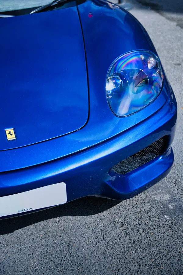 Détail de l'avant d'une Ferrari bleue métallisée, l'emblème du cheval cabré sur le côté de l'aile, et une partie de la prise d'air du pare-chocs.