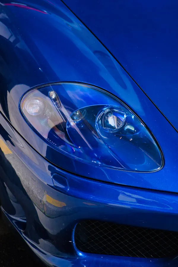 Gros plan sur le phare avant d'une Ferrari 360 Modena bleu métallisé, montrant le détail de l'optique transparente avec projecteur xénon, intégré dans la carrosserie aérodynamique brillante.