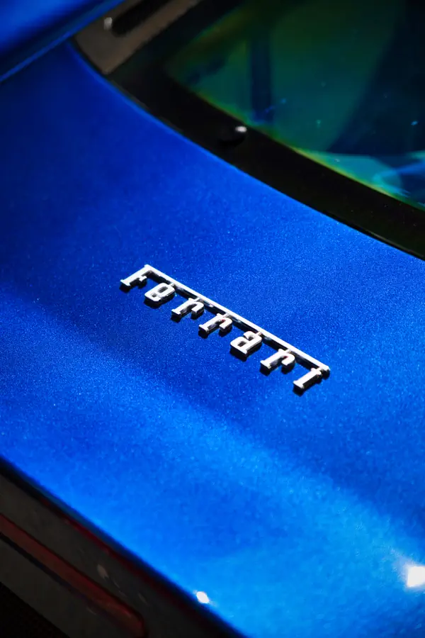 Badge Ferrari en lettres chromées sur la carrosserie bleu métallisé d'une supercar, montrant le détail de la peinture brillante avec ses reflets et une partie du pare-brise visible en haut de l'image.