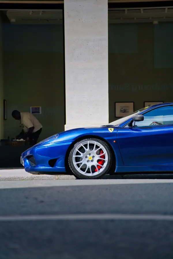 Ferrari 360 Challenge Stradale bleue vue de profil, stationnée devant un bâtiment moderne avec colonne en béton, montrant sa jante en alliage argentée à rayons multiples, son étrier de frein rouge visible, et l'emblème Ferrari sur l'aile avant.