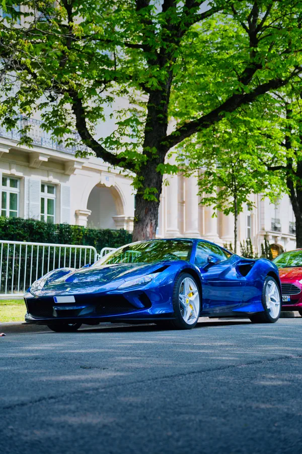 Ferrari F8 Tributo bleu électrique stationnée dans une rue parisienne, avec un grand arbre feuillu au printemps et un élégant immeuble haussmannien blanc en arrière-plan, montrant les lignes aérodynamiques de la supercar et ses jantes en alliage argentées avec étriers jaunes.