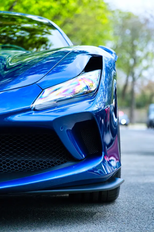 Gros plan sur l'avant d'une Ferrari F8 Tributo bleu électrique, montrant le design agressif du pare-chocs avec ses grandes prises d'air latérales, le phare LED effilé et la calandre en nid d'abeille, photographiée en extérieur avec un fond de verdure printanière.
