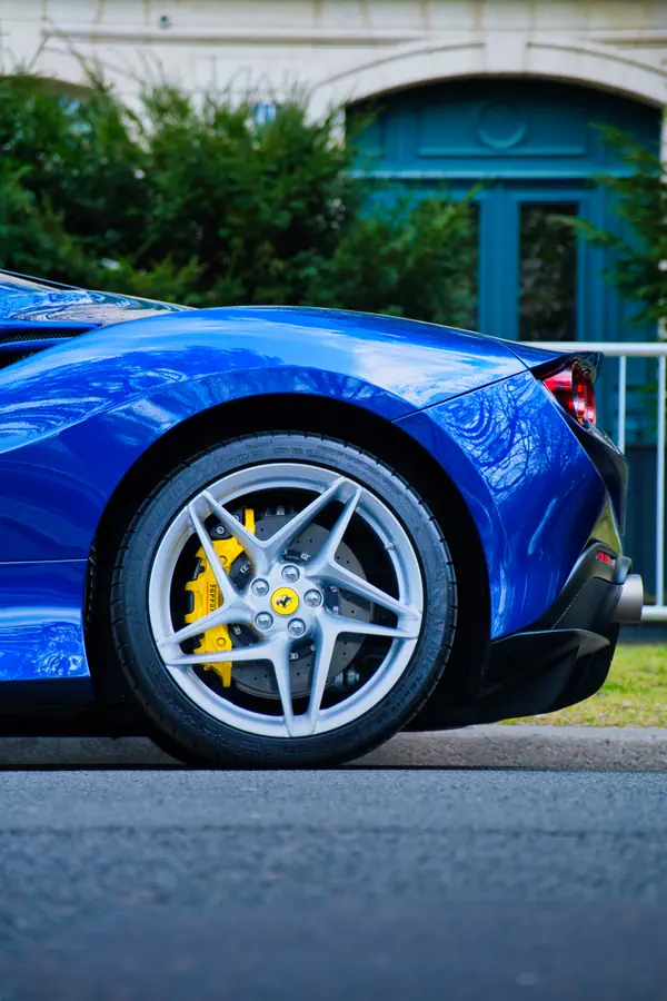 Détail de la roue arrière d'une Ferrari F8 Tributo bleue, montrant sa jante en alliage à cinq branches en étoile, son étrier de frein jaune Ferrari, son disque de frein perforé et un aperçu du feu arrière, avec en arrière-plan un bâtiment ancien à porte bleue entouré de végétation.