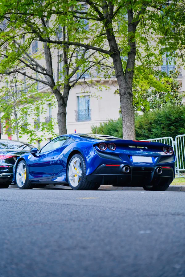 Ferrari F8 Tributo bleu électrique photographiée de trois-quarts arrière, stationnée dans une rue parisienne au printemps, avec ses feux arrière ronds caractéristiques, ses doubles sorties d'échappement, ses jantes en alliage argentées et des arbres feuillus et immeubles haussmanniens en arrière-plan.