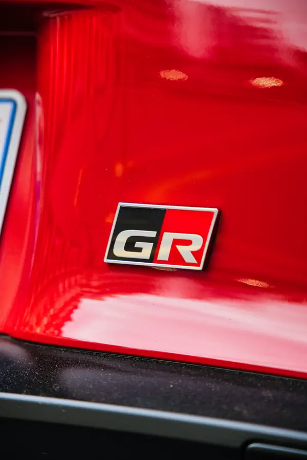 Badge GR (Gazoo Racing) sur la carrosserie rouge brillante d'une Toyota, montrant l'emblème rectangulaire divisé en noir et rouge avec les lettres "GR" en blanc, photographié en gros plan avec des reflets sur la peinture.