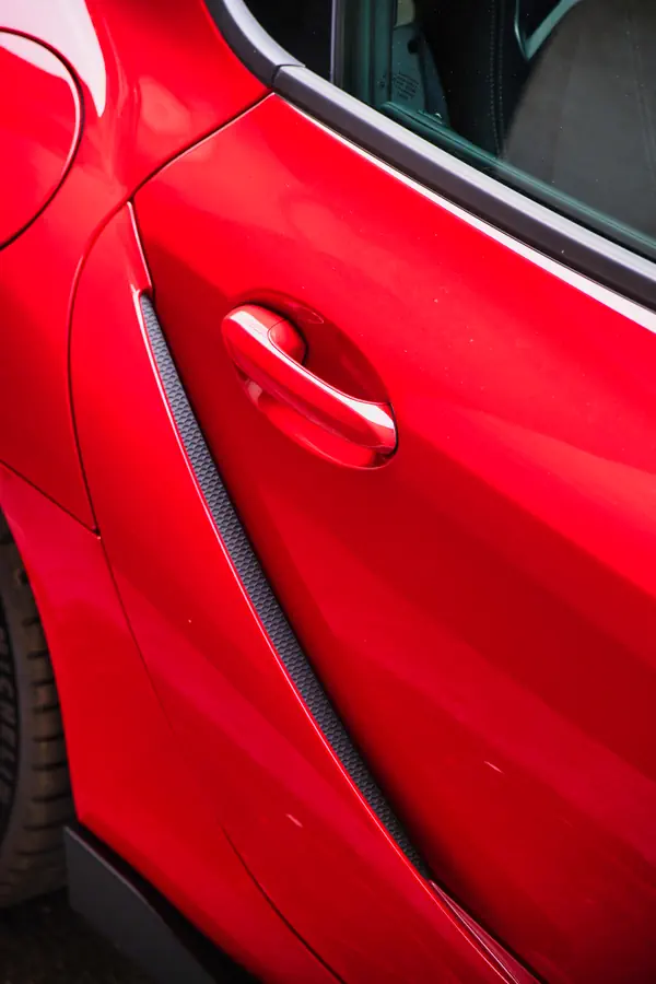 Détail de la portière d'une Toyota GR Yaris rouge, montrant la poignée de porte intégrée avec finition chromée, une bande décorative en fibre de carbone texturée le long du bas de la portière, et la ligne de carrosserie sportive avec son reflet brillant sur la peinture rouge vif.