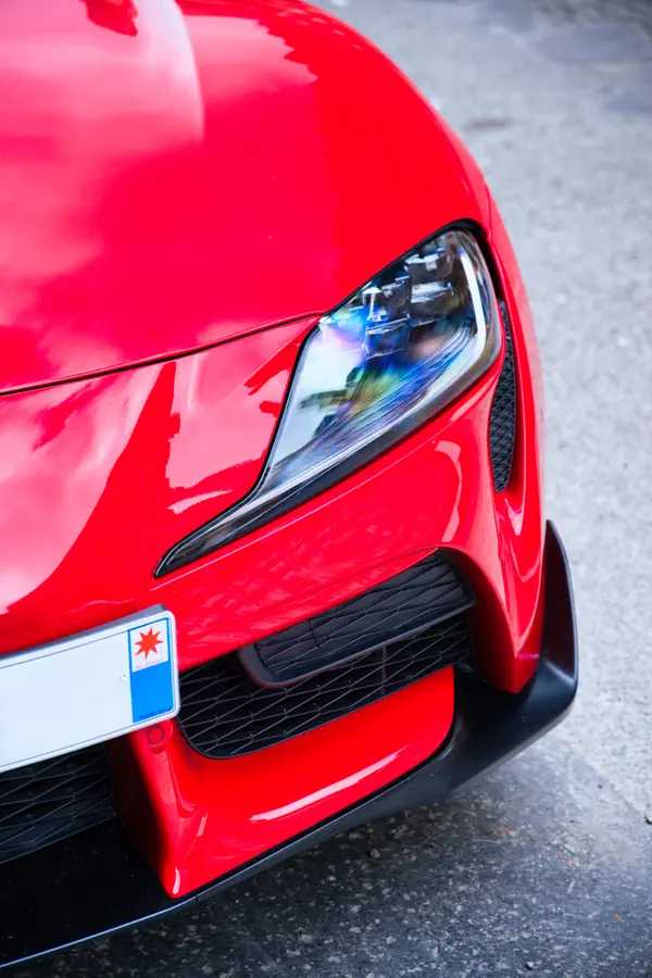 Gros plan sur l'avant d'une Toyota GR Supra rouge, montrant son phare LED effilé, sa large prise d'air latérale avec grille noire, une partie de la plaque d'immatriculation, et le pare-chocs aérodynamique avec splitter noir.