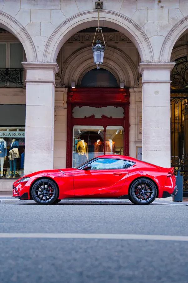 Toyota GR Supra rouge vue de profil, stationnée devant une boutique de luxe avec une entrée ornée d'arches en pierre et d'une porte rouge élégante, montrant la silhouette sportive du coupé avec ses lignes fluides, ses jantes noires et sa posture basse et athlétique.