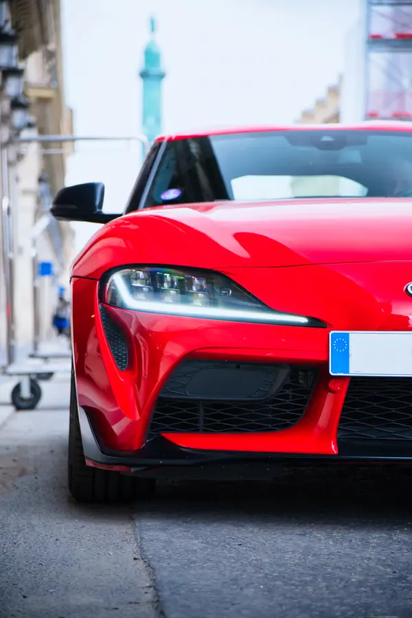 Avant d'une Toyota GR Supra rouge vif photographiée dans une rue parisienne, montrant son phare LED distinctif avec signature lumineuse, sa large calandre en nid d'abeille, sa plaque d'immatriculation européenne, et la colonne Vendôme visible en arrière-plan flou.