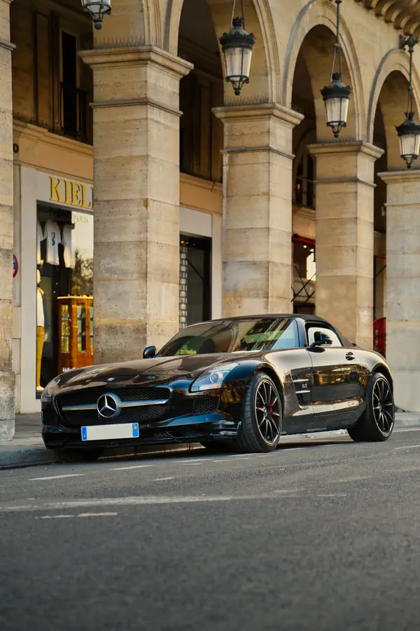 Mercedes-Benz SLS AMG noire stationnée devant les arcades de la rue de Rivoli, avec ses colonnes en pierre et lanternes caractéristiques, montrant la silhouette élégante du roadster avec sa calandre à étoile proéminente, ses jantes multi-branches et ses étriers de frein rouges.