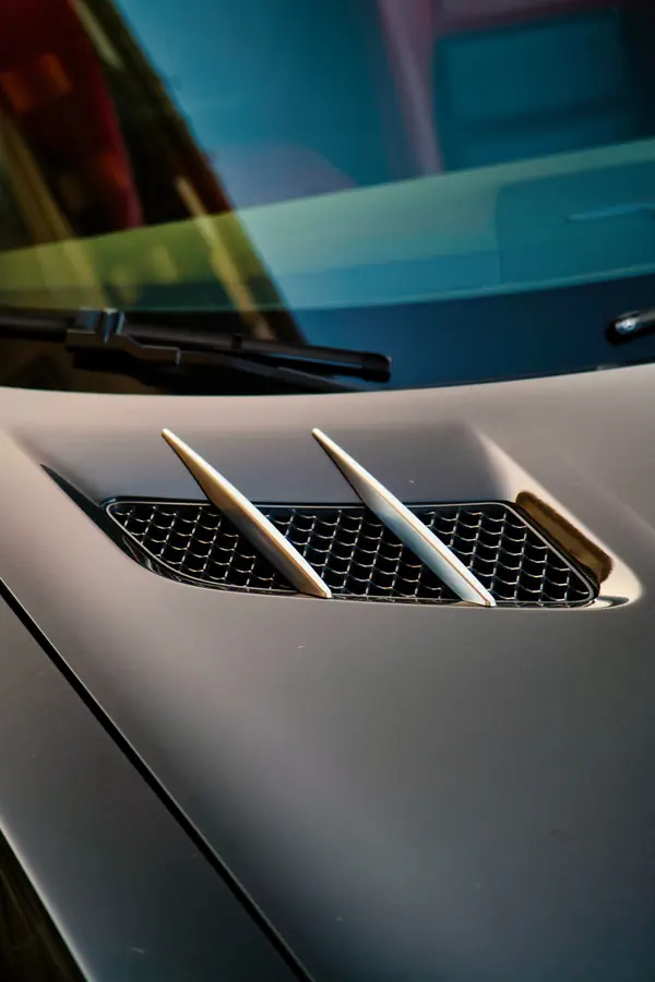 Détail d'une ouïe d'aération latérale sur l'aile d'une Mercedes-Benz SLS AMG grise, montrant trois lamelles chromées traversant une grille en nid d'abeille noire.