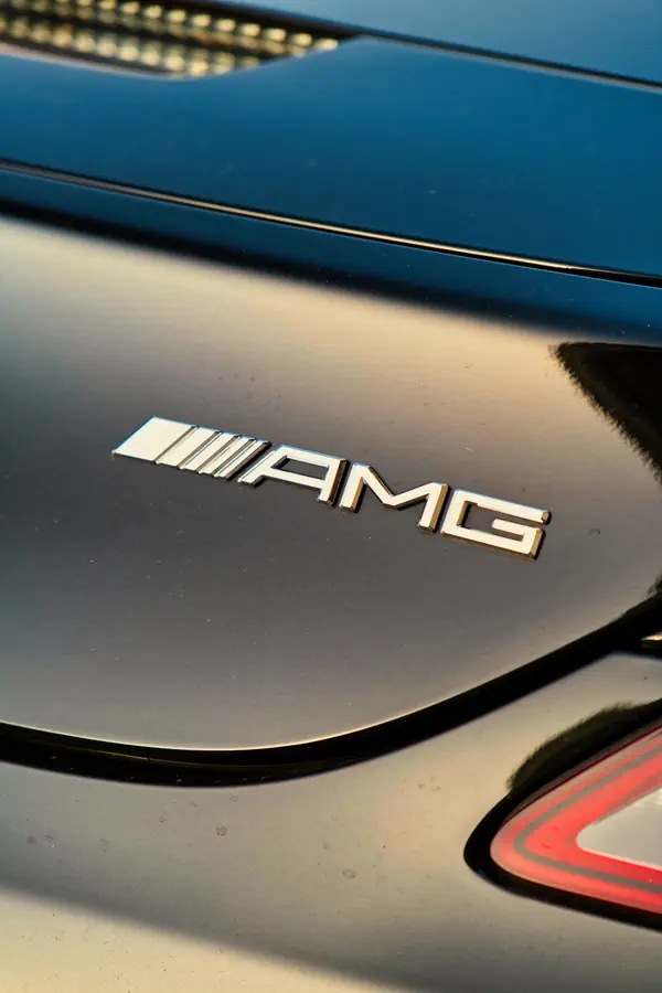 Badge AMG chromé en relief sur la carrosserie grise d'une Mercedes-Benz, photographié en gros plan avec une partie du feu arrière visible en bas à droite et des reflets sur la peinture métallisée.