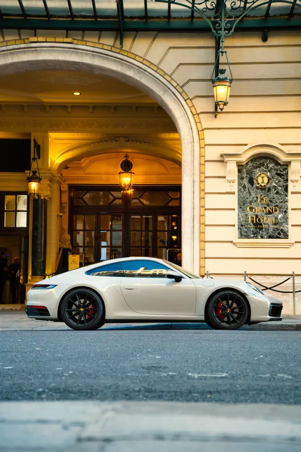 Porsche 911 blanche vue de profil, stationnée devant l'entrée élégante du Grand Hôtel de Paris avec son arche en pierre, ses lanternes dorées et sa plaque en marbre vert.