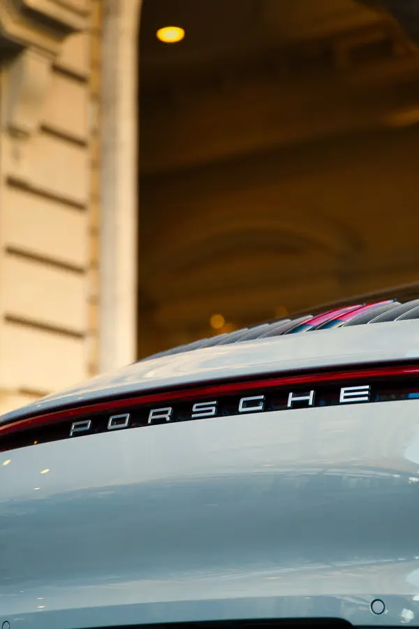 Détail de l'arrière d'une Porsche 911 blanche montrant le lettrage "PORSCHE" chromé sur la bande lumineuse rouge reliant les feux arrière, avec un bâtiment parisien en pierre beige partiellement visible en arrière-plan.