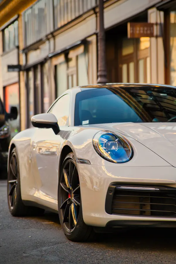 Avant d'une Porsche 911 blanche photographiée en angle, stationnée dans une rue commerçante parisienne, montrant son phare rond emblématique à LED, sa jante sportive avec étrier de frein rouge.