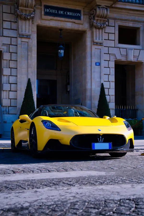 Maserati MC20 jaune vif stationnée devant l'entrée majestueuse de l'Hôtel de Crillon à Paris, montrant la supercar avec sa calandre distinctive ornée du trident Maserati, ses phares LED effilés et sa carrosserie aérodynamique, encadrée par deux topiaires coniques et la façade en pierre de taille du palace historique.