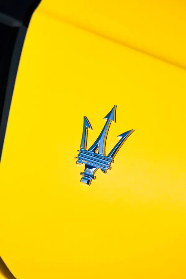 Emblème Maserati chromé représentant le trident de Neptune en relief sur la carrosserie jaune vif d'une Maserati MC20, photographié en gros plan avec un contraste saisissant entre le métal bleuté et le fond éclatant.