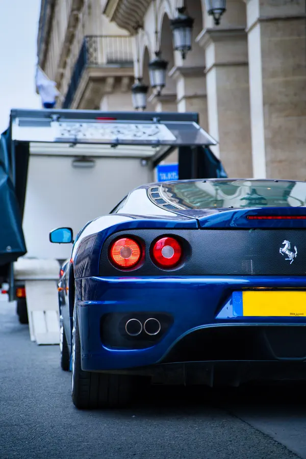 Arrière d'une Ferrari 360 Modena bleu métallisé stationnée dans une rue parisienne, montrant ses feux arrière ronds caractéristiques allumés, son double échappement et l'emblème du cheval cabré, avec la façade d'un bâtiment historique en pierre en arrière-plan.