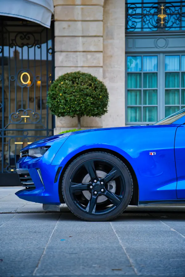 Détail de l'avant d'une Chevrolet Camaro bleu électrique stationnée devant l'entrée d'un hôtel parisien de luxe, montrant sa roue avant avec jante noire à cinq branches.