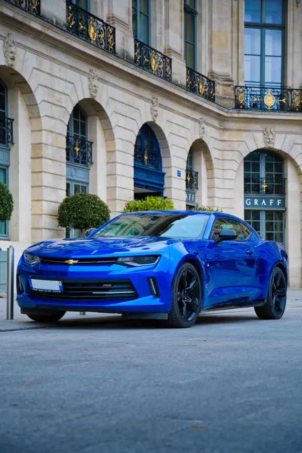 Chevrolet Camaro bleu électrique photographiée de trois-quarts avant sur la place Vendôme à Paris, devant la boutique Graff, montrant son logo Chevrolet doré et ses jantes noires, avec en arrière-plan la façade néoclassique ornée de balcons en fer forgé doré et de buis taillés.