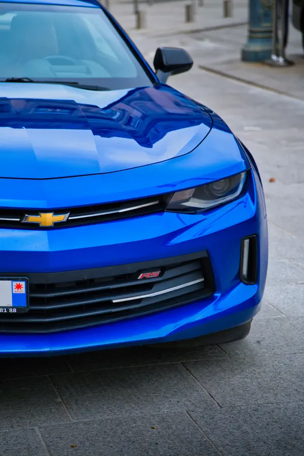 Gros plan sur l'avant d'une Chevrolet Camaro RS bleu électrique, montrant le détail du phare LED, le logo Chevrolet doré en forme de nœud papillon sur la calandre noire et le badge RS rouge sur le pare-chocs inférieur.
