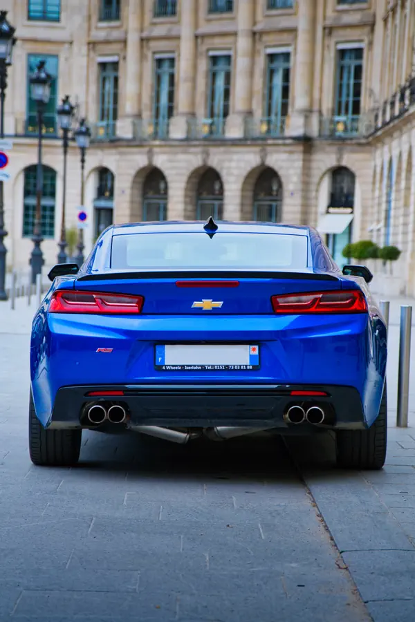 Arrière d'une Chevrolet Camaro RS bleu électrique photographiée sur la place Vendôme à Paris, montrant ses feux arrière LED caractéristiques, le logo Chevrolet doré, le badge RS rouge, ses quatre sorties d'échappement et sa silhouette musclée, avec en arrière-plan la façade classique d'un bâtiment historique avec ses arcades et lanternes.