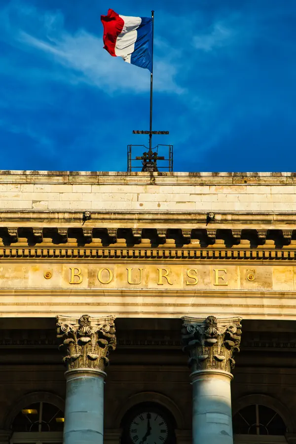 Façade néoclassique du Palais Brongniart (La Bourse de Paris) avec son inscription dorée "BOURSE", ses colonnes corinthiennes et son fronton, surmonté du drapeau tricolore français flottant sur un ciel bleu.