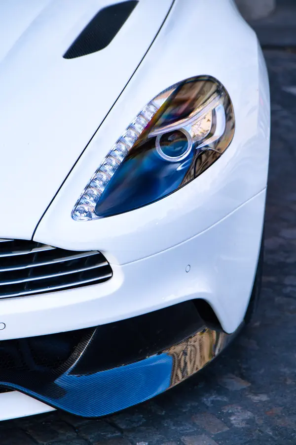 Gros plan sur le phare avant d'une Aston Martin Vanquish blanche, montrant le détail de l'optique avec sa signature lumineuse LED distinctive, photographiés sur une surface pavée.