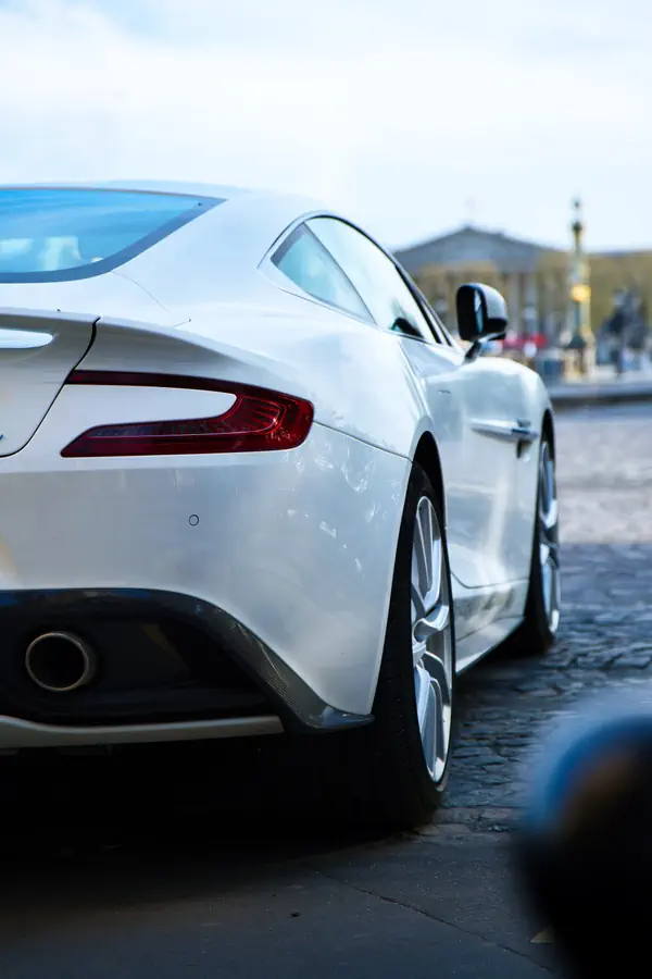 Vue arrière d'une élégante voiture de sport blanche Aston Martin garée sur des pavés de la Place de la Concorde à Paris, avec l'assemblée nationale en arrière-plan.