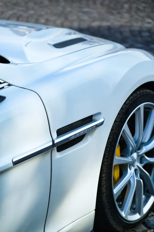 Gros plan sur l'aile avant d'une Aston Martin blanche montrant les détails de l'évent latéral chromé, la carrosserie sculptée et une jante en alliage argentée avec étrier de frein jaune.