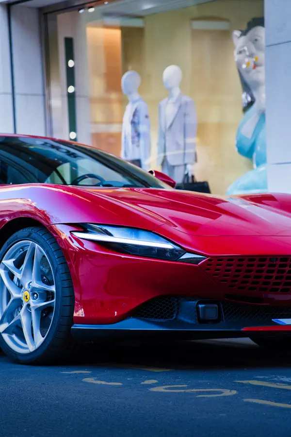Ferrari Roma rouge éclatante garée devant une boutique de luxe, montrant le design élégant de sa partie avant avec ses phares effilés, sa calandre distinctive et ses jantes en alliage à cinq branches.