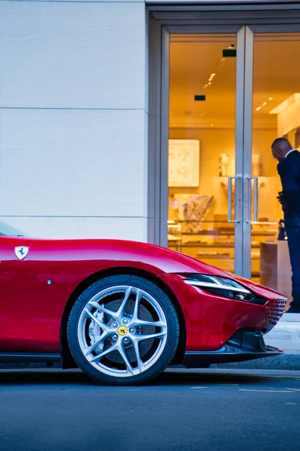Partie avant d'une Ferrari Roma rouge stationnée devant une boutique de luxe, mettant en valeur l'emblème du cheval cabré, les jantes en alliage complexes et les phares LED distinctifs, avec l'entrée élégamment éclairée du magasin en arrière-plan.