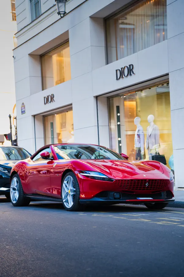 Ferrari Roma décapotable rouge éclatante stationnée devant la boutique Dior à Paris, mettant en valeur ses lignes élégantes, sa calandre distinctive et ses jantes en alliage, créant un contraste saisissant avec la façade blanche du bâtiment de luxe.