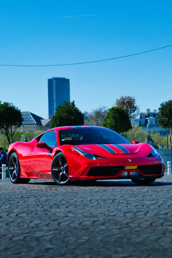 Ferrari 458 Speciale rouge vif avec bandes noires sur le capot, photographiée sur une place pavée à Paris par une journée ensoleillée, avec la Tour Montparnasse visible en arrière-plan et un ciel bleu éclatant.
