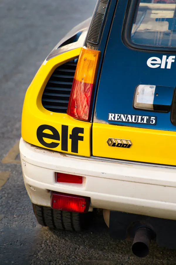 Détail de l'arrière d'une Renault 5 Turbo aux couleurs Elf, montrant le feu arrière caractéristique, la prise d'air latérale jaune, le logo Renault 5, l'emblème Elf et le badge Codi sur la carrosserie bleu et jaune, avec l'échappement visible sous le pare-chocs blanc.