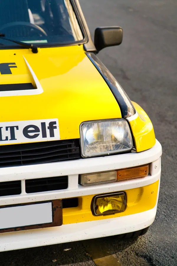 Gros plan sur l'avant d'une Renault 5 Turbo aux couleurs Elf, montrant le phare rectangulaire caractéristique, le clignotant, le phare antibrouillard jaune et le pare-chocs blanc contrastant avec la carrosserie jaune vif ornée du logo Elf noir.