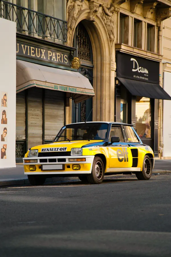 Renault 5 Turbo aux couleurs Elf jaune et bleue stationnée devant la boutique "Au Vieux Paris" dans une rue parisienne historique.