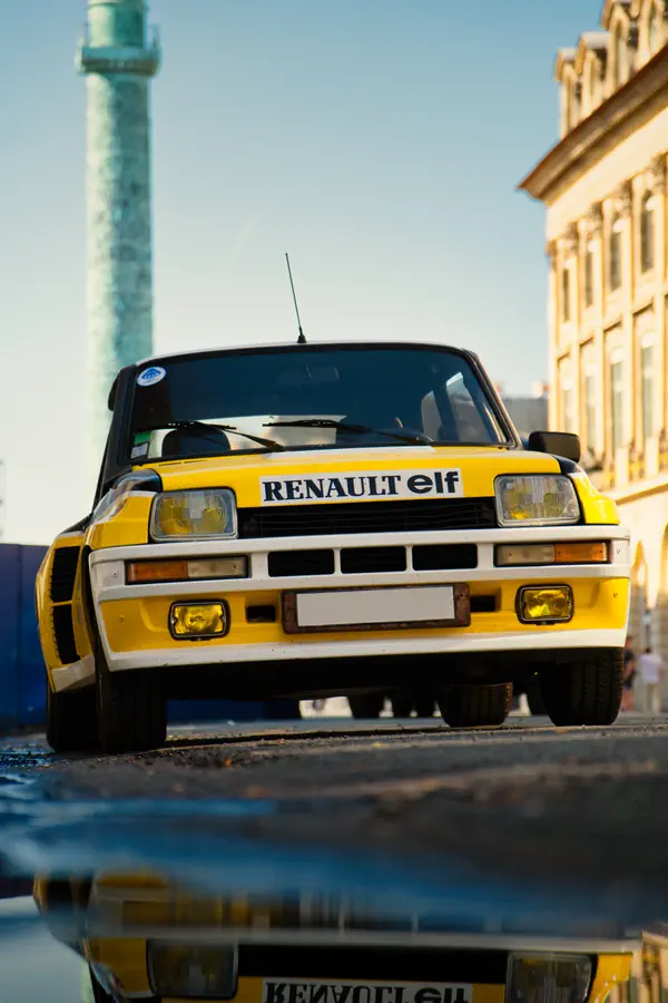 Vue frontale d'une Renault 5 Turbo aux couleurs Elf photographiée à Paris, avec la colonne de la place Vendôme visible en arrière-plan. La voiture jaune et noire emblématique est capturée au ras du sol, créant un reflet partiel dans une flaque d'eau au premier plan.