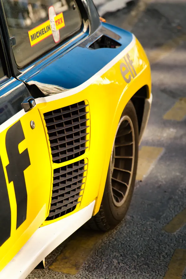 Gros plan sur l'aile élargie d'une Renault 5 Turbo aux couleurs Elf, montrant la prise d'air latérale caractéristique avec sa grille noire, la jante spécifique, et un autocollant Michelin TRX visible sur la vitre.