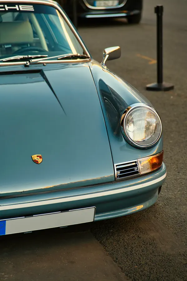 Détail de l'avant d'une Porsche 911 classique bleu clair métallisé, montrant le phare rond caractéristique, le clignotant rectangulaire et l'emblème Porsche sur l'aile.