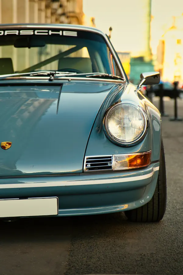 Gros plan sur l'avant d'une Porsche 911 classique bleu ciel métallisé, capturant le phare rond caractéristique, le clignotant rectangulaire et l'écusson Porsche sur l'aile. Le pare-brise arbore le lettrage "PORSCHE" et la lumière dorée du coucher de soleil se reflète sur la carrosserie.