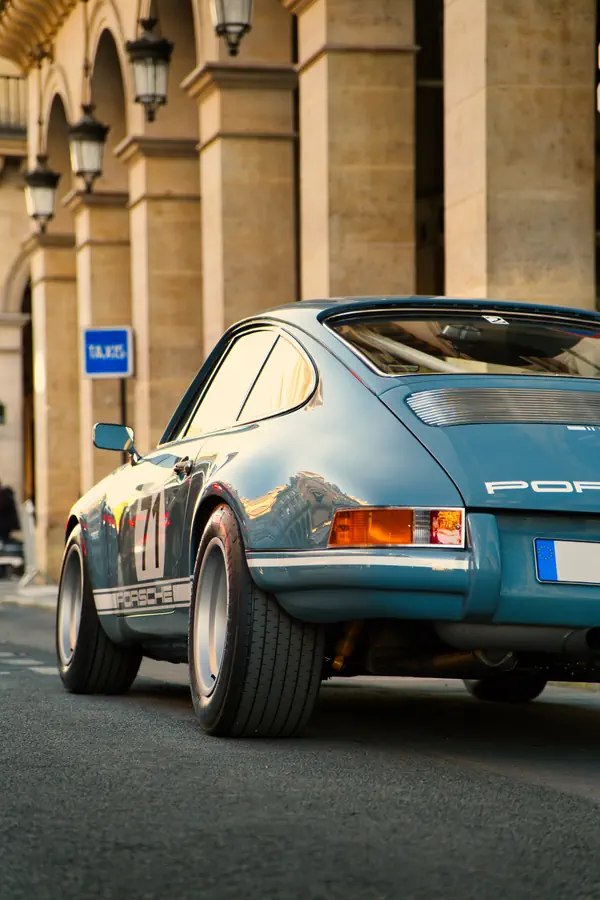 Vue arrière d'une Porsche 911 classique bleu ciel avec le numéro 71 et des bandes Porsche sur les flancs, photographiée devant un bâtiment historique parisien en pierre.