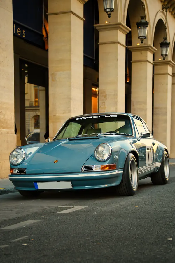Porsche 911 classique bleu ciel avec le numéro 71 et bandes latérales Porsche, photographiée devant les arcades élégantes de la Place Vendôme à Paris.
