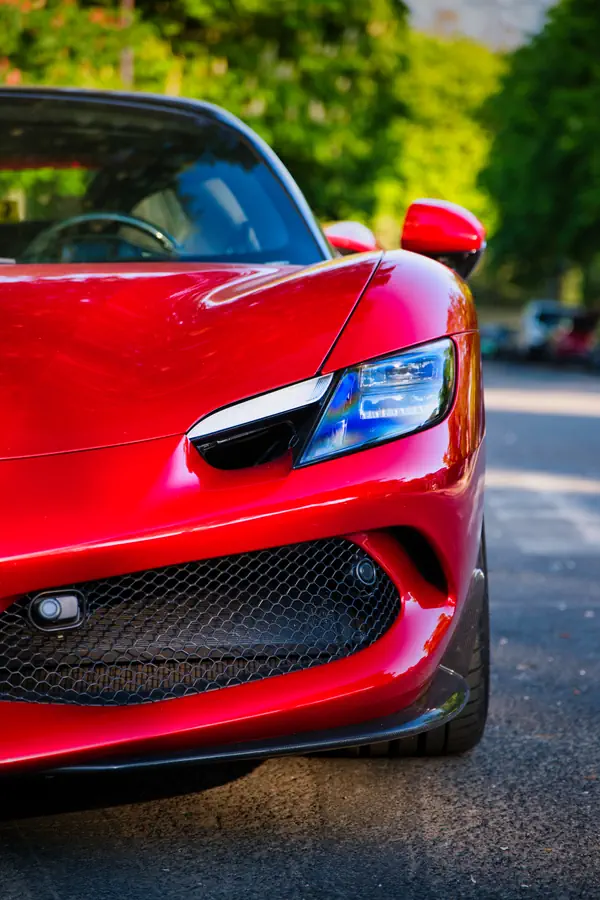 Gros plan sur le phare avant et la calandre d'une Ferrari 296 GTB rouge éclatant, montrant le design agressif du phare LED, la grille en nid d'abeille noire et les lignes aérodynamiques sculptées de la carrosserie.